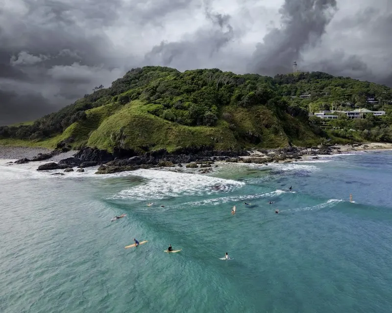 Surfers in the water off The Pass under a stormy sky, with the green headland behind